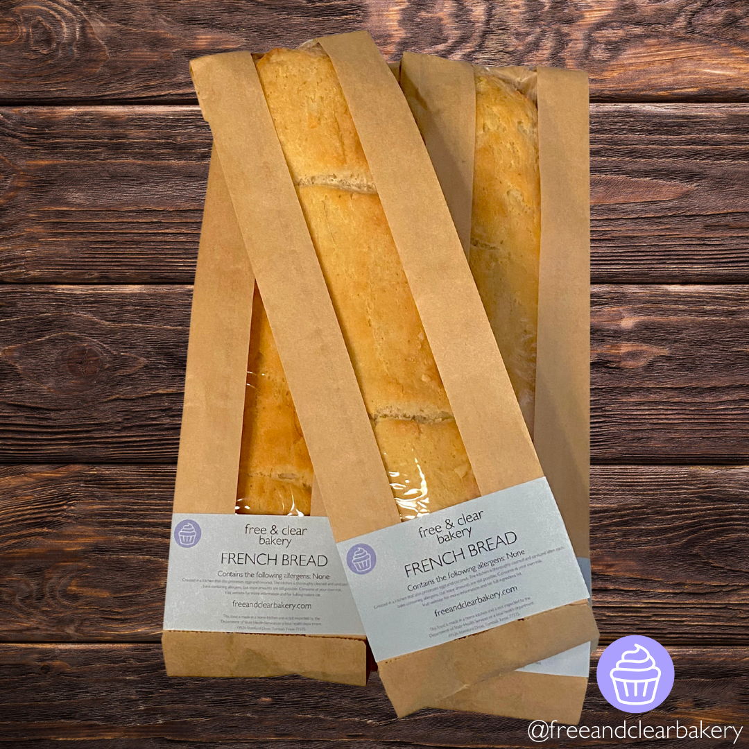 Overhead shot of two baguettes parallel to each other with a third lying diagonally on top of them. All baguettes are perfectly golden brown inside brown paper bags with clear plastic windows running the length. Each bag has a Free & Clear Bakery rectangular label on the bottom listing ingredients and the website.