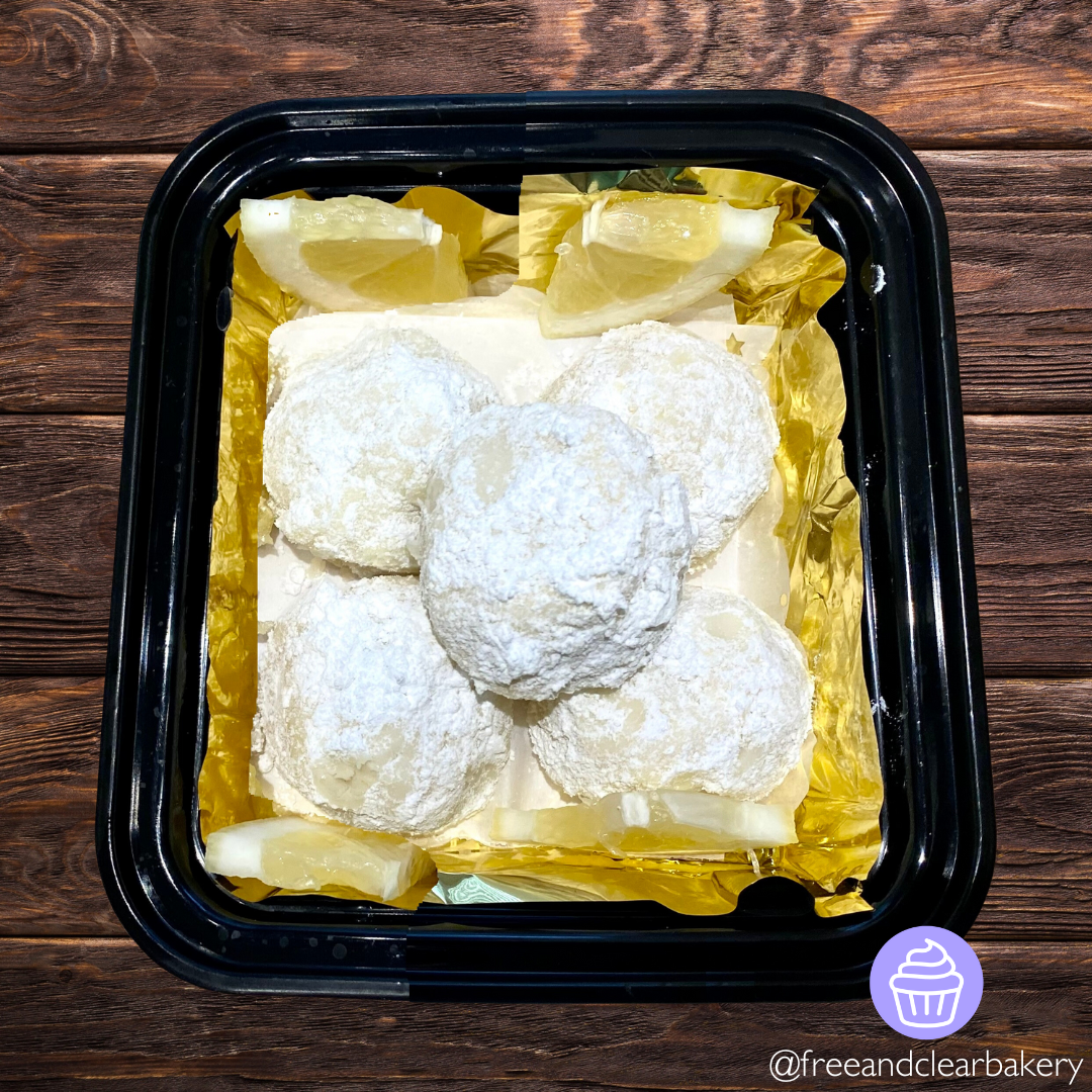 Black food container on top of a dark brown wood table. In the container is five round balls of lemony dough, rolled in powder sugar, on top of gold foil, with lemon slice wedges as decoration in the corners.