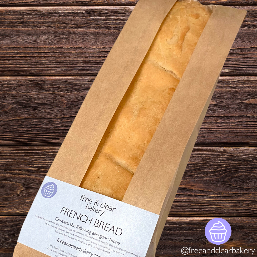 One single golden brown baguette packaged in a brown paper bag with a clear window running lengthwise. At the bottom of the loaf is a rectangular Free & Clear Bakery label with the logo, "French Bread", and ingredient information.