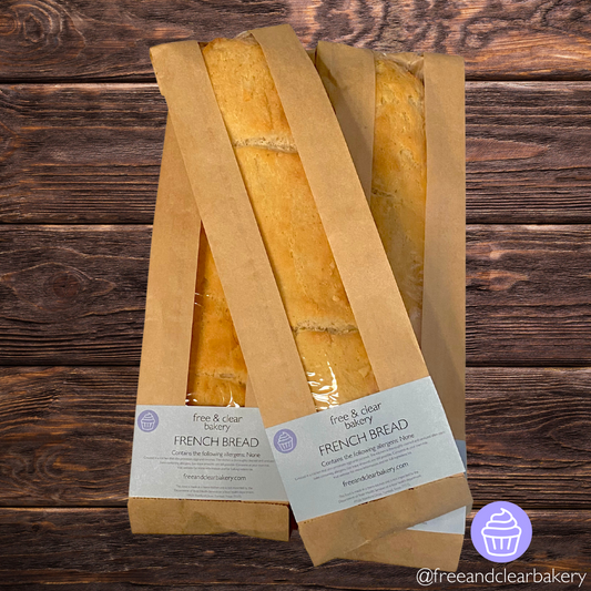 Overhead shot of two baguettes parallel to each other with a third lying diagonally on top of them. All baguettes are perfectly golden brown inside brown paper bags with clear plastic windows running the length. Each bag has a Free & Clear Bakery rectangular label on the bottom listing ingredients and the website. 