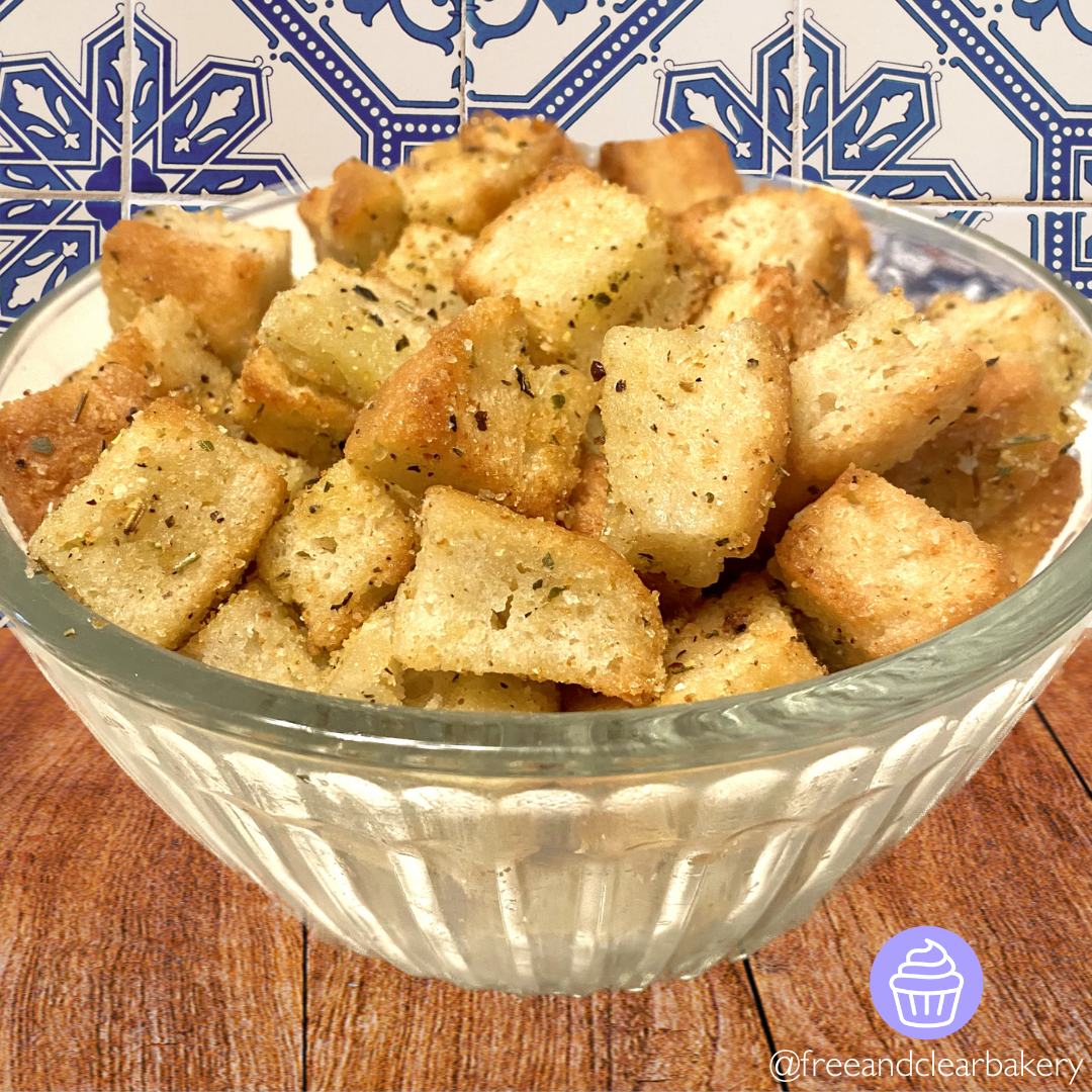 Close up image of beautifully crispy baked croutons in a clear glass bowl, on top of a wooden table with a blue mosaic tile wall behind.