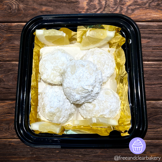 Black food container on top of a dark brown wood table. In the container is five round balls of lemony dough, rolled in powder sugar, on top of gold foil, with lemon slice wedges as decoration in the corners. 