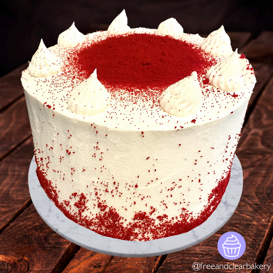 Giant red velvet cake with white frosting and Red Velvet crumbs as decoration around the sides and also in a circle on the top. The top has white frosting dollops in a circle as a border.