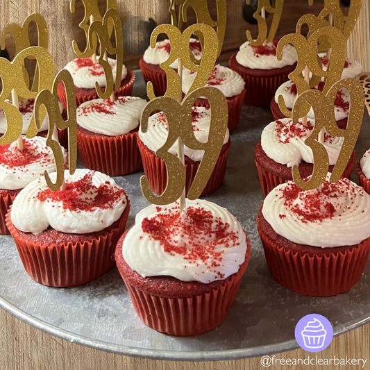 Red velvet cupcakes with red cupcake liners, white frosting, and red velvet crumbs sprinkled on top. Each cupcake has a gold number 39 decoration on a stick inserted into each cupcake for a 39th birthday party